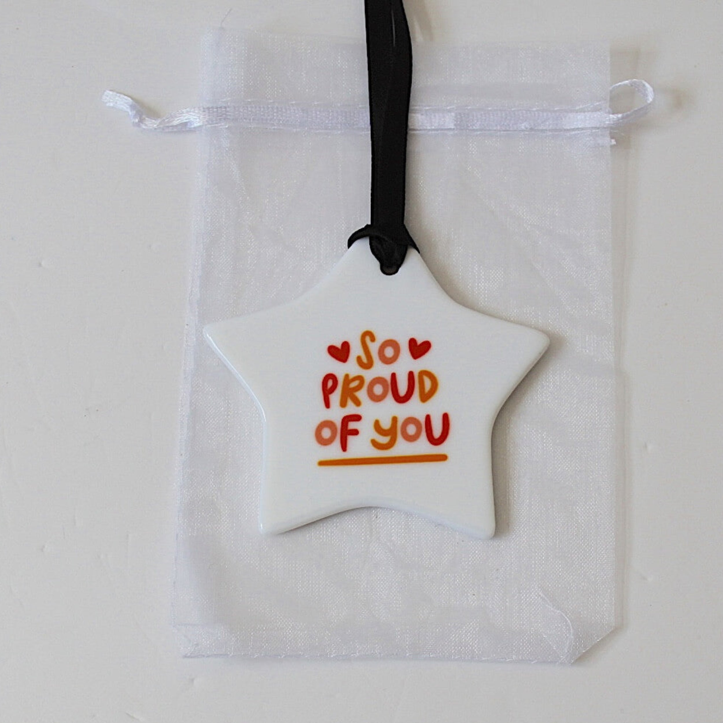 A white star-shaped ornament with the words 'SO PROUD OF YOU' in red and orange lettering, hanging from a black ribbon over a white drawstring pouch on a light background.