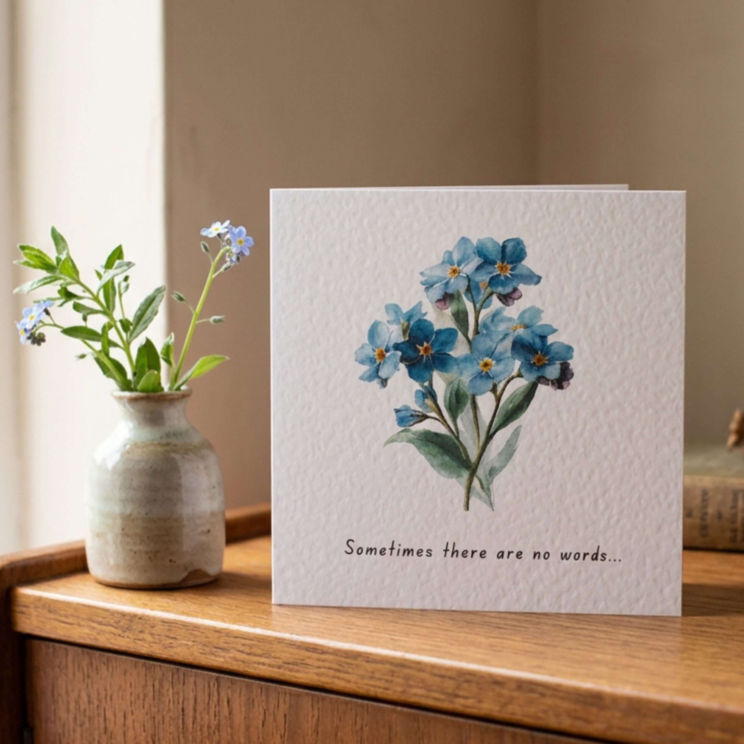 A square greeting card with a watercolor illustration of blue forget-me-nots and the printed phrase 'Sometimes there are no words.
