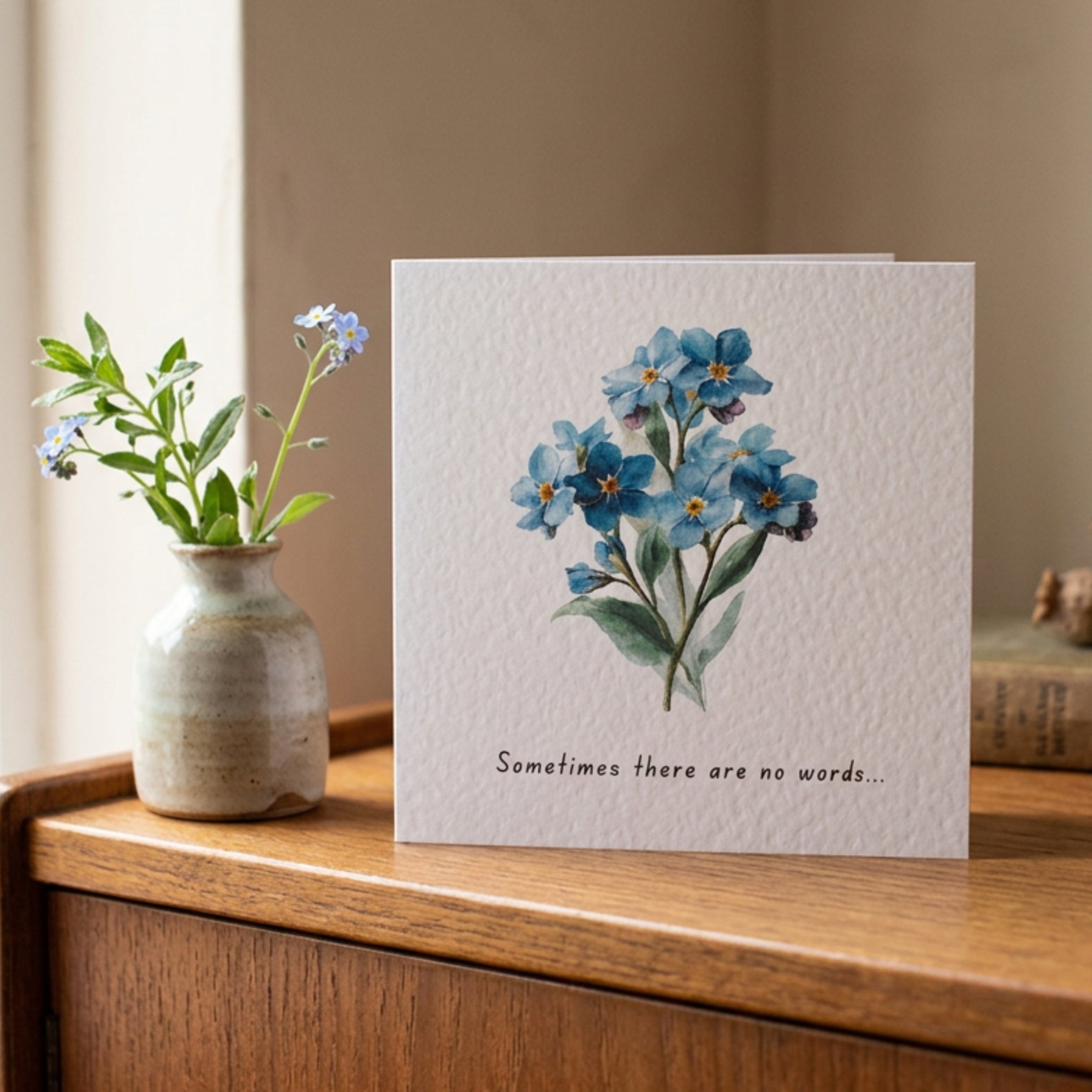 A square greeting card with a watercolor illustration of blue forget-me-nots and the printed phrase 'Sometimes there are no words.