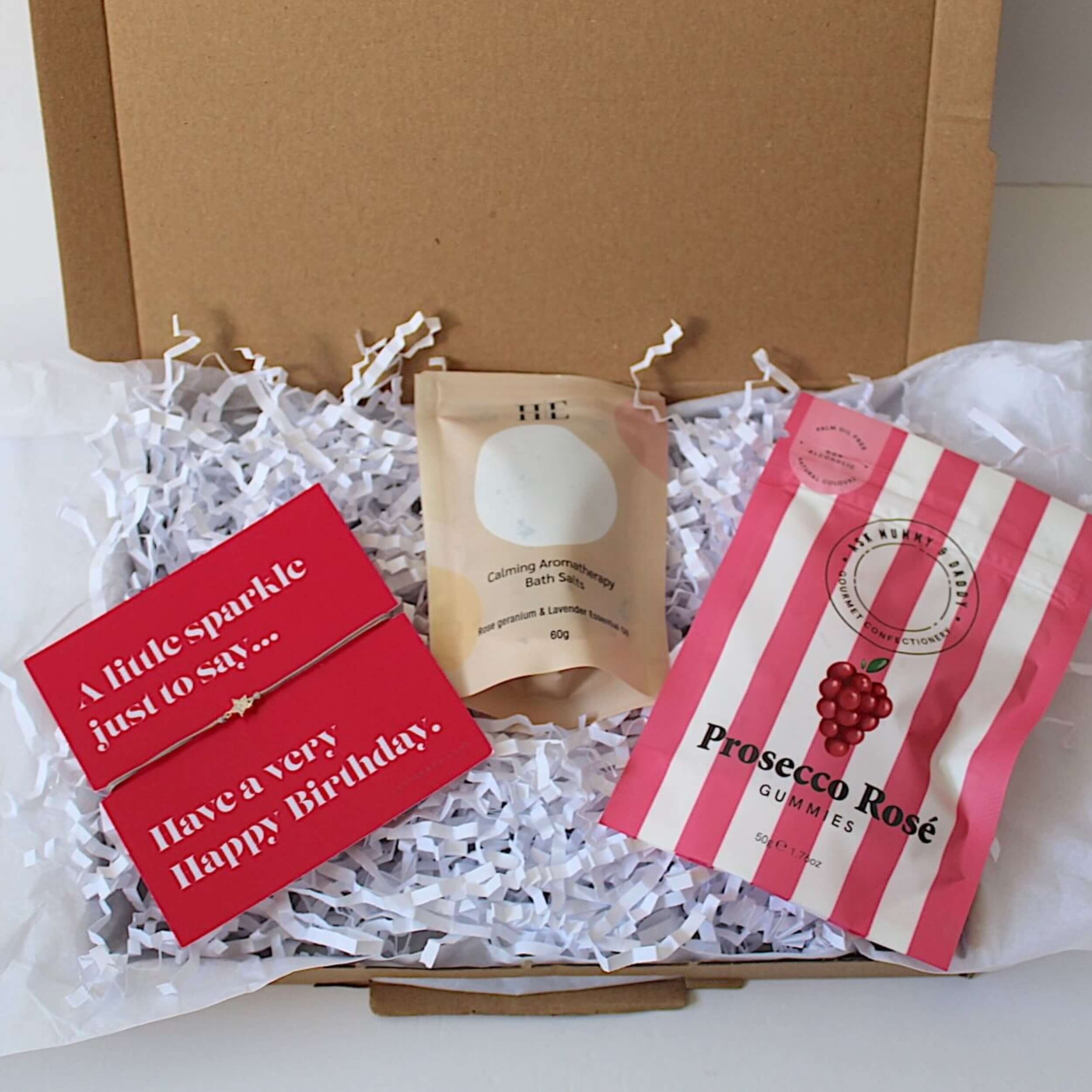 A curated gift box set containing a aromatherapy bath salts, a pink and white striped pouch of Prosecco Rosé, a red greeting card, with a bracelet with star  all arranged in white crinkle paper inside an open gift box. 