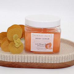 Sunday Rain Mango Butter Coconut Oil Scrub in clear jar with tropical flowers on wooden tray.