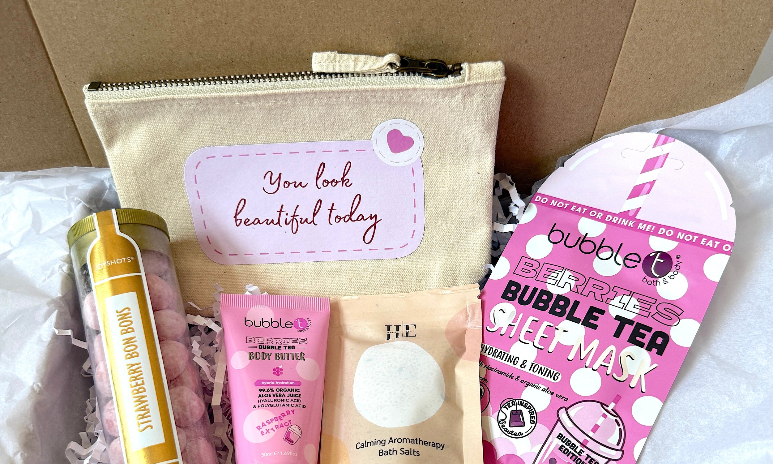 A curated gift box set containing a beige canvas pouch with a pink label reading 'You look beautiful today', raspberry body butter, 60g aromatherapy bath salts, berries bubble tea sheet mask, and a tube of strawberry bom boms,  all arranged in white crinkle paper inside a brown cardboard box. 