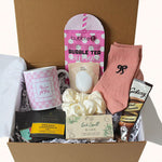 A curated gift box set in a brown cardboard box, featuring a pink checkered mug, a pink bubble tea sheet mask, a peach-colored sock with a black bow, a cream scrunchie, a box of Taylors organic chamomile tea, and a chocolate bar.