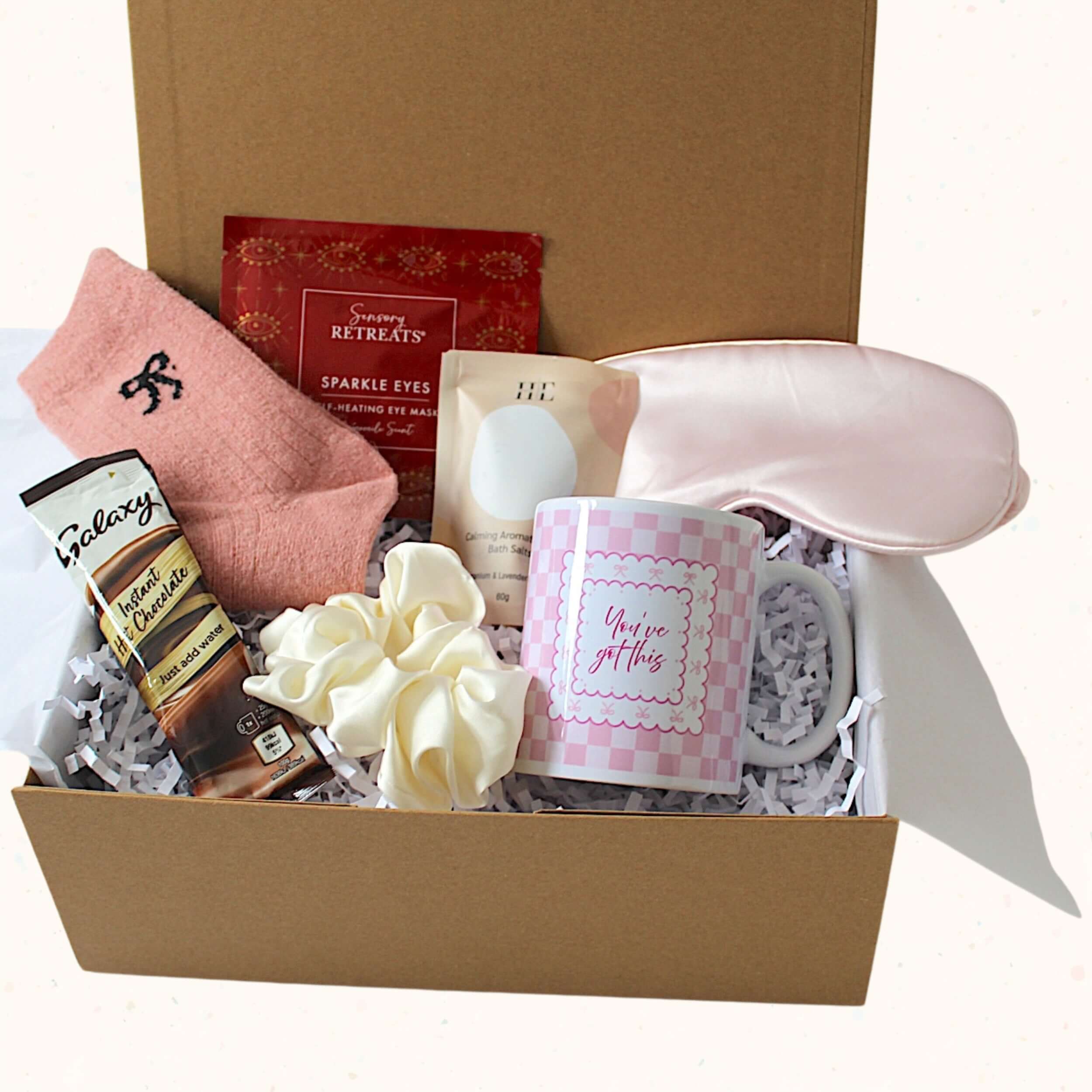 A curated gift box set in a brown cardboard box filled with white paper shred, featuring a pink towel sock, a Galaxy chocolate bar, a pink and white checkered mug that says 'You're got this', a cream hair scrunchie, a pink satin sleep mask, a small bottle, and a red packaged treat.
