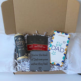 An open cardboard gift box filled with white tissue paper and crinkle paper shred, containing a can of Barista Coffee Co., a box of Taylor's Breakfast Tea, a gray square gift card with a message for Dad, and a Joybars 'Best Dad' chocolate bar.