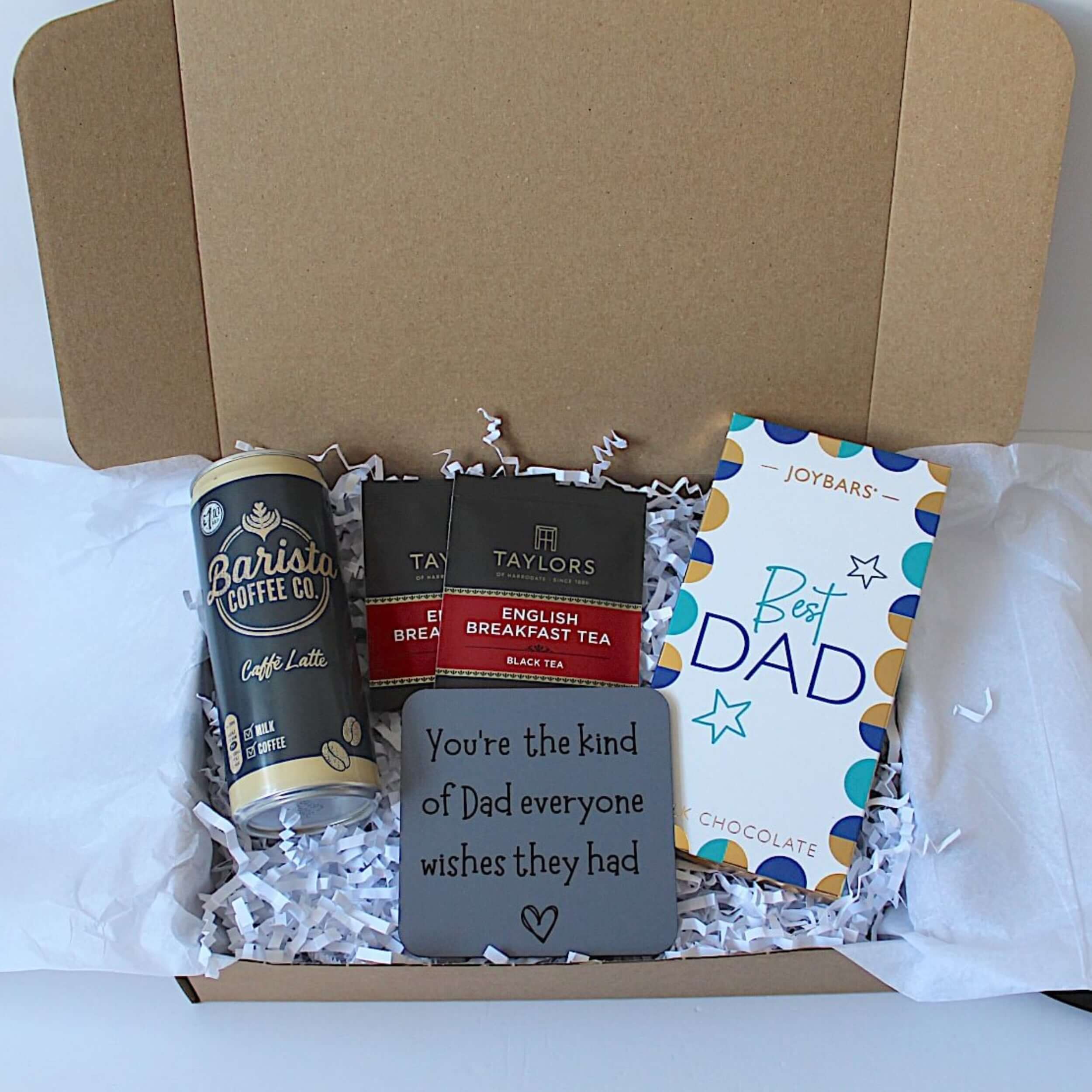 An open cardboard gift box filled with white tissue paper and crinkle paper shred, containing a can of Barista Coffee Co., a box of Taylor's Breakfast Tea, a gray square gift card with a message for Dad, and a Joybars 'Best Dad' chocolate bar.
