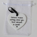 A white ceramic heart-shaped ornament with black printed text reading 'Distance means nothing when you have someone so special!' and a small black heart at the bottom, hanging from a black ribbon. It is displayed inside a clear white drawstring pouch on a light background.