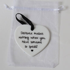 A white ceramic heart-shaped ornament with black printed text reading 'Distance means nothing when you have someone so special!' and a small black heart at the bottom, hanging from a black ribbon. It is displayed inside a clear white drawstring pouch on a light background.