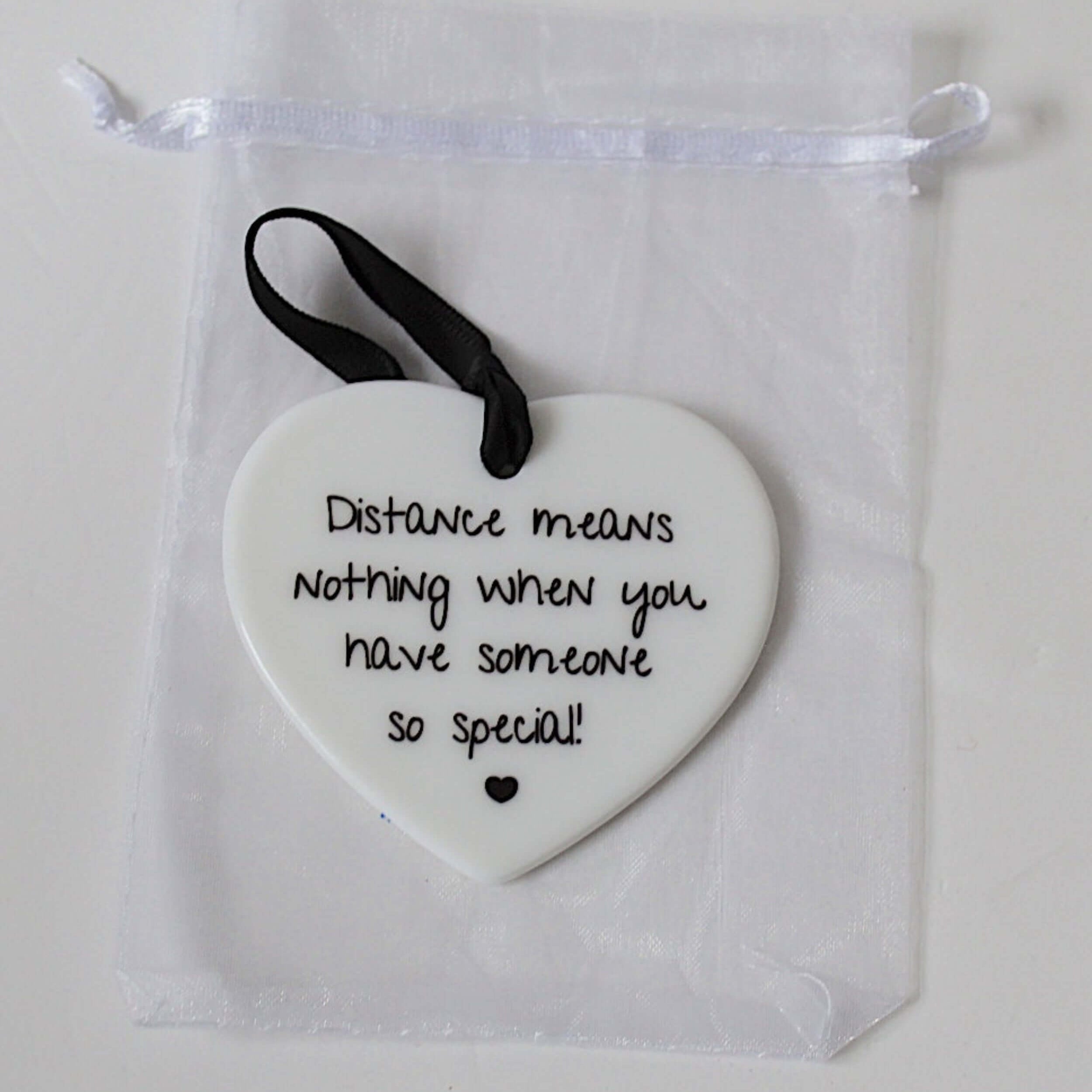 A white ceramic heart-shaped ornament with black printed text reading 'Distance means nothing when you have someone so special!' and a small black heart at the bottom, hanging from a black ribbon. It is displayed inside a clear white drawstring pouch on a light background.