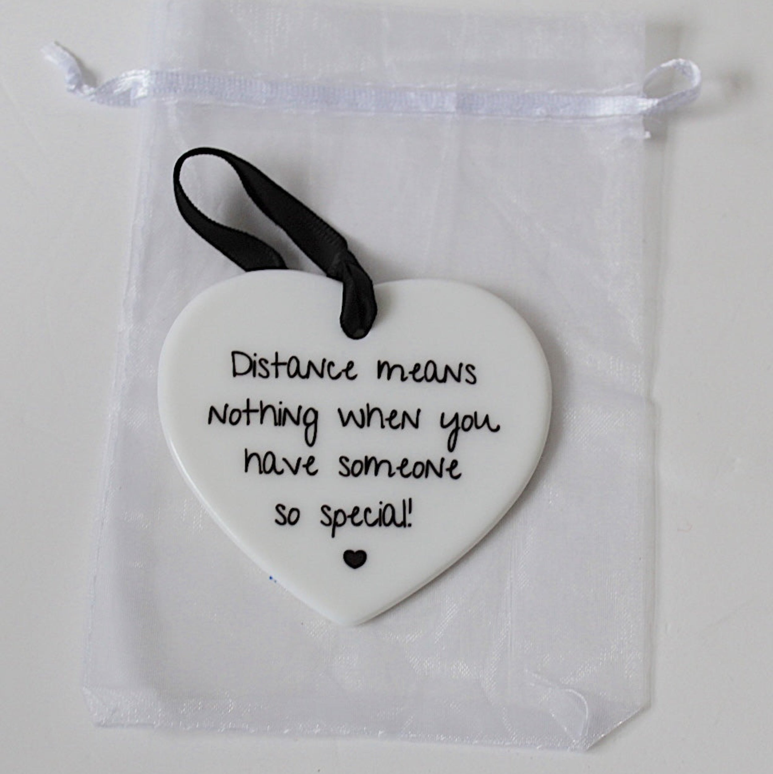 A white ceramic heart-shaped ornament with black printed text reading 'Distance means nothing when you have someone so special!' and a small black heart at the bottom, hanging from a black ribbon. It is displayed inside a clear white drawstring pouch on a light background.