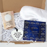 A curated gift box set from Serenity Retreats, featuring a white satin sleep mask, a blue Luna Eyes self-healing eye mask box, a beige bath soak pouch, and a white heart-shaped ornament nestled in white shredded paper.