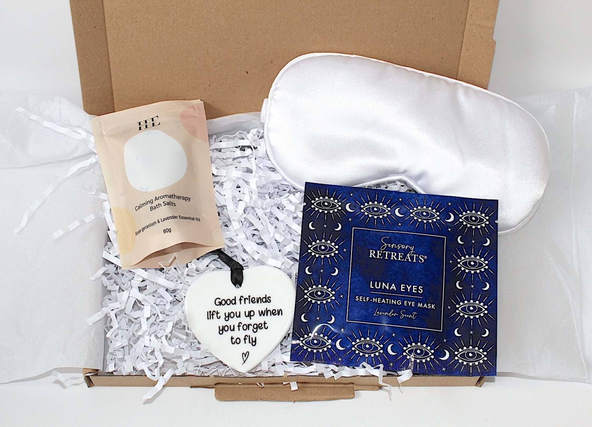 A curated gift box set from Serenity Retreats, featuring a white satin sleep mask, a blue Luna Eyes self-healing eye mask box, a beige bath soak pouch, and a white heart-shaped ornament nestled in white shredded paper.