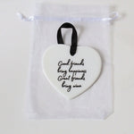 A white heart-shaped ceramic ornament with black cursive text reading 'Good friends bring happiness, Great friends bring wine,' hanging from a black ribbon loop inside a clear drawstring pouch.