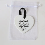 A white heart-shaped ceramic ornament with black cursive text reading 'Good friends bring happiness, Great friends bring wine,' hanging from a black ribbon loop inside a clear drawstring pouch.