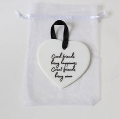A white heart-shaped ceramic ornament with black cursive text reading 'Good friends bring happiness, Great friends bring wine,' hanging from a black ribbon loop inside a clear drawstring pouch.