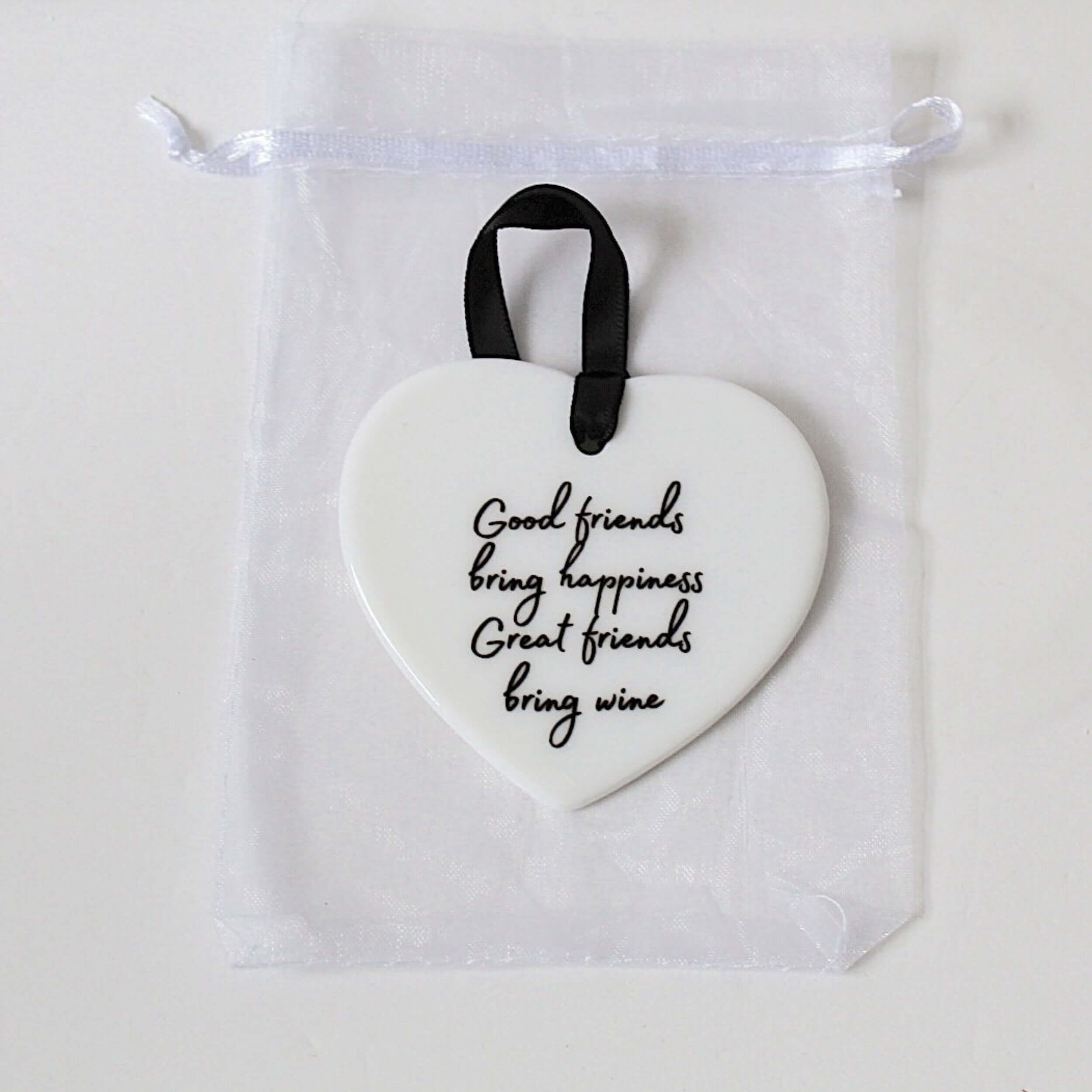 A white heart-shaped ceramic ornament with black cursive text reading 'Good friends bring happiness, Great friends bring wine,' hanging from a black ribbon loop inside a clear drawstring pouch.