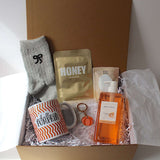A curated gift box set in a brown magnetic closure gift box filled with shredded paper, containing a grey sock with a black bow, a white mug with orange wavy stripes and 'hello PUMPKIN' text, a gold honey sheet face mask, sunday rain mango and coconut body wash, aromatherapy bath salts  and a small orange pumpkin keychain. 