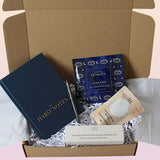 An open kraft gift box filled with self-care and stationery items, including a navy blue Habit Notes journal, a blue Luna Etes eye mask pack, a beige sheet mask packet, and a white eye cream tube on white crinkle paper.