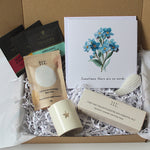 A curated gift box set from Taylor's Organics, featuring a cream ceramic candle with a star, a boxed essential oil, 3 soy wax tea lights bath salts, tea bags, and a floral greeting card nestled in white paper shred inside a brown cardboard box.