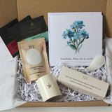 A curated gift box set from Taylor's Organics, featuring a cream ceramic candle with a star, a boxed essential oil, 3 soy wax tea lights bath salts, tea bags, and a floral greeting card nestled in white paper shred inside a brown cardboard box.