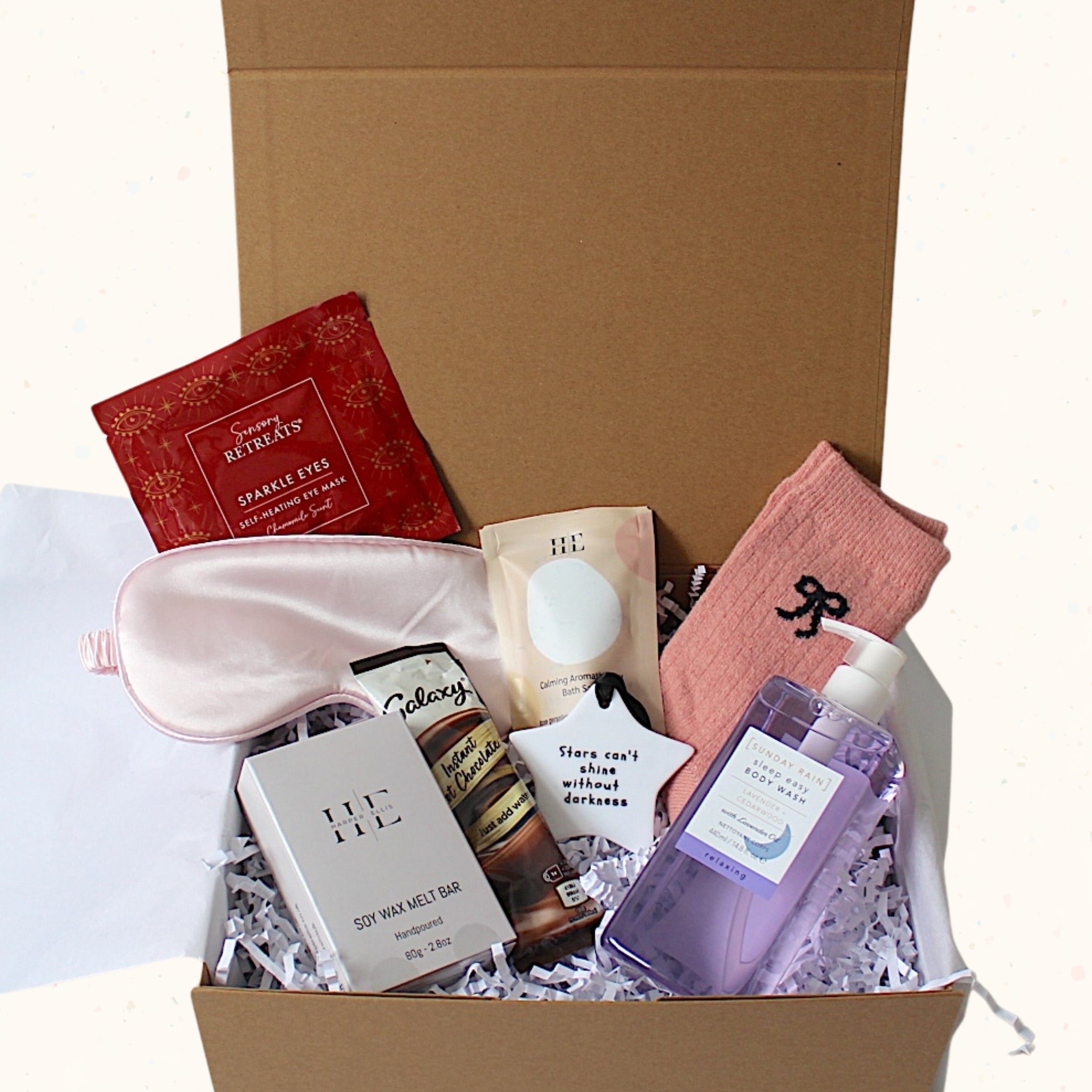 A curated gift box set in a brown cardboard box filled with shreds of paper. The set includes a pink satin sleep mask, a red sparkly eye mask packet, a pink sock with a black bow, a purple hand sanitizer bottle, a white body wax melt bag, and a small brown bottle.