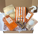 A curated gift box set of personal care products arranged in a cardboard box filled with white paper shred. The set includes an orange body wash bottle, a gold honey nourishing pouch, an orange and white striped Italian Spritz Gummies pouch, a beige sheet mask packet, and a brown hair scrunchie.