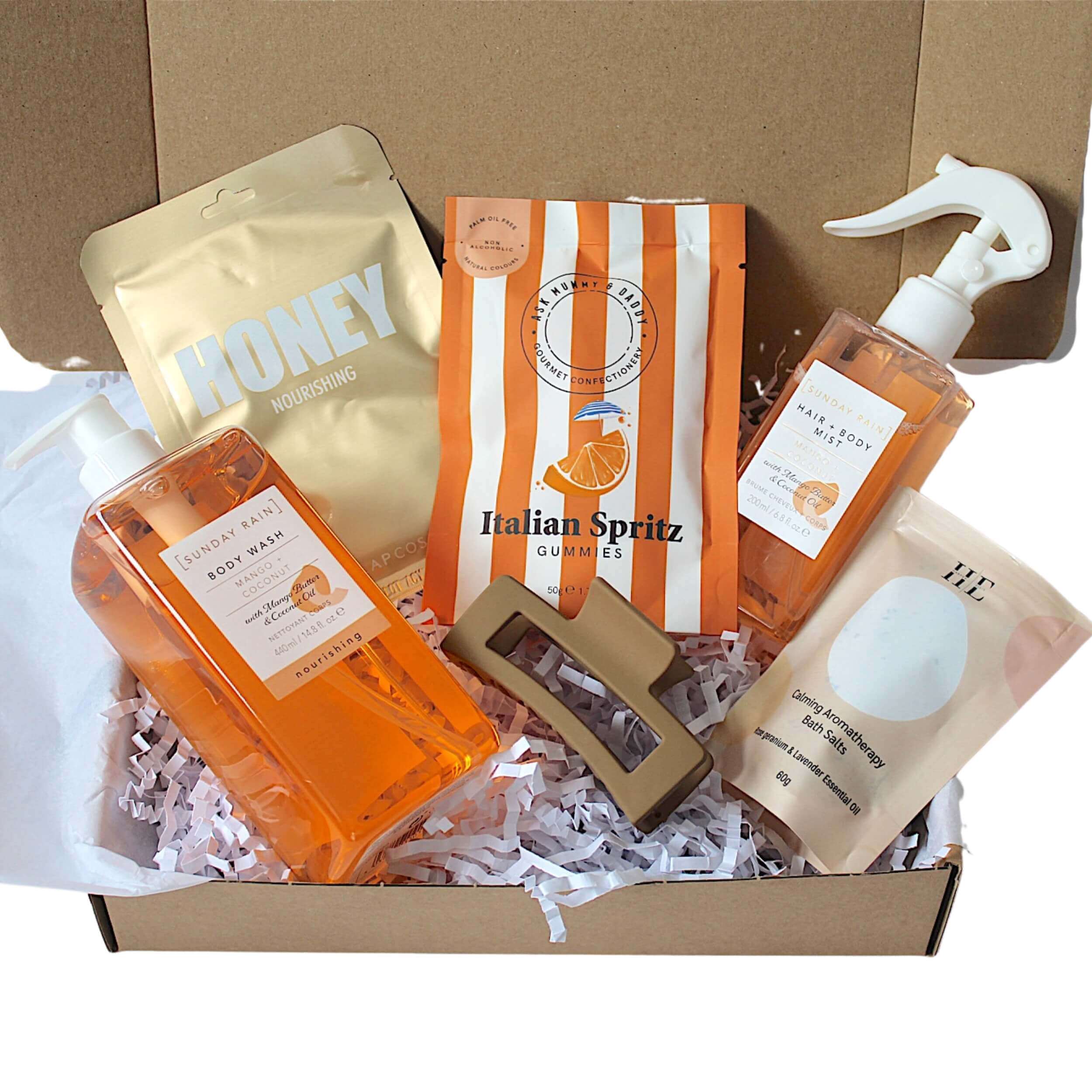 A curated gift box set of personal care products arranged in a cardboard box filled with white paper shred. The set includes an orange body wash bottle, a gold honey nourishing pouch, an orange and white striped Italian Spritz Gummies pouch, a beige sheet mask packet, and a brown hair scrunchie.