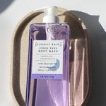 Purple bottle of Sunday Rain body wash on a textured beige surface