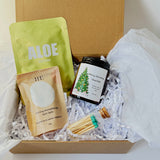 A gift box filled with bath and beauty items on shredded white paper. Visible items include a green pouch labeled ALOE, a peach-colored packet labeled Grooming Aromatherapy Bath Salts, a small black jar labeled Merry Christmas Charity, and a glass vial with teal-tipped applicators.