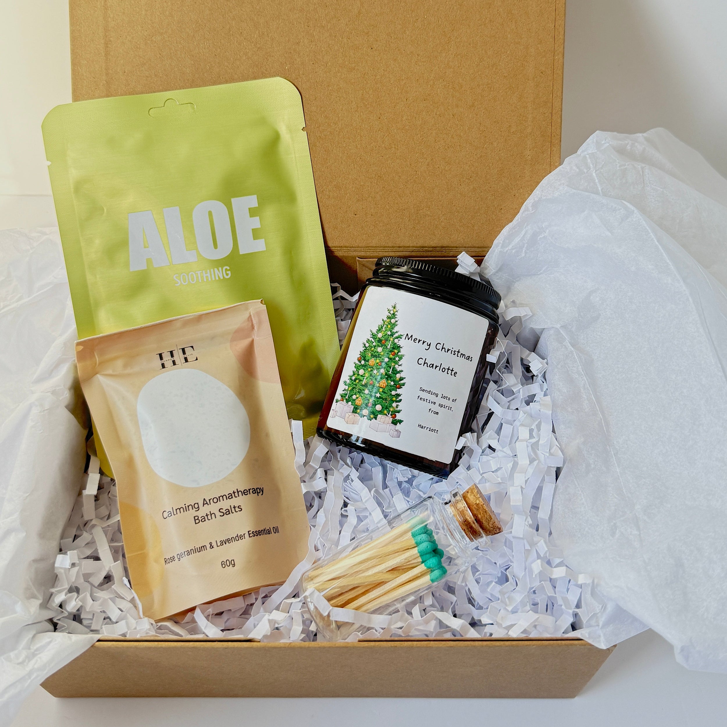 A gift box filled with bath and beauty items on shredded white paper. Visible items include a green pouch labeled ALOE, a peach-colored packet labeled Grooming Aromatherapy Bath Salts, a small black jar labeled Merry Christmas Charity, and a glass vial with teal-tipped applicators.